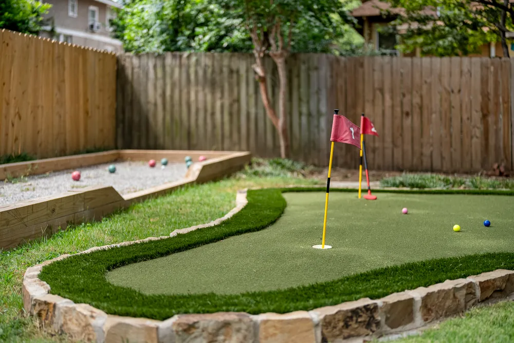 The backyard putting green with two flags, and the bocce court behind it along a wooden fence.